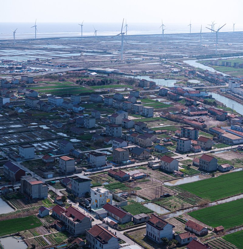 Aerial view of a low-rise residential settlement with agricultural plots and wind turbines on the horizon