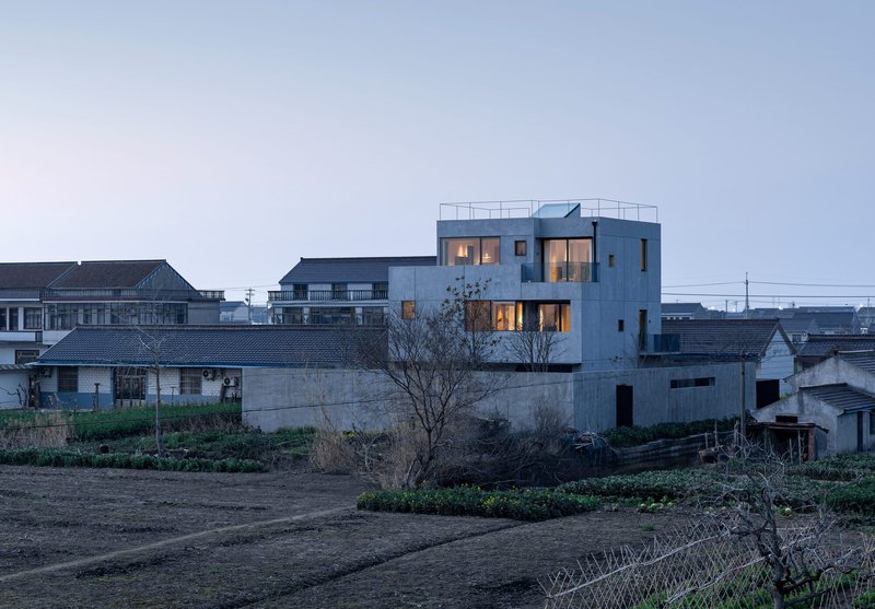 White concrete house with rooftop terrace rising above neighboring buildings and cultivated fields at dusk