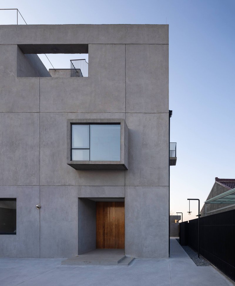 Street-facing concrete facade with timber entry door and projecting box window under a clear sky