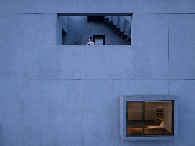 Concrete panel facade with recessed upper balcony where a figure stands beside an interior staircase