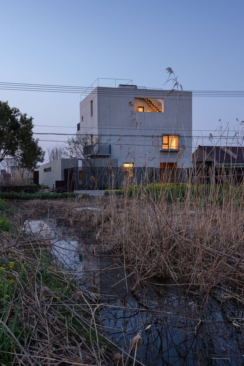 Concrete facade with lit windows at dusk across a narrow irrigation channel with dry winter grasses