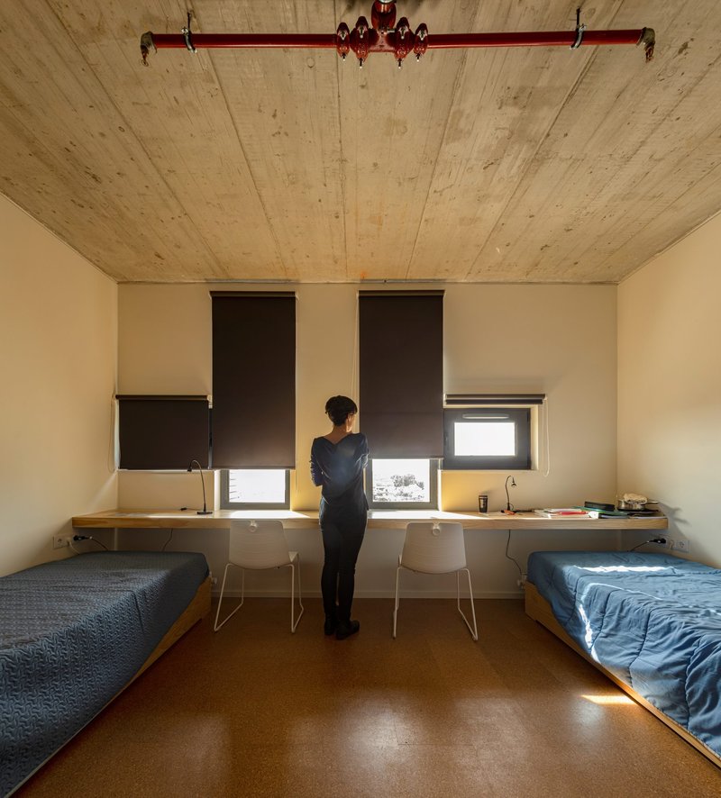 Shared bedroom with two beds and floating desk beneath partially drawn roller shades with a person standing
