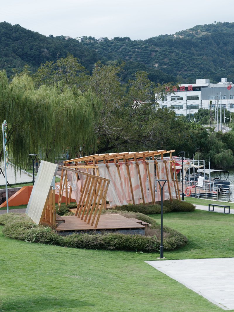 Angled view of the tilting pavilion structure with exposed timber beams in a lakeside park setting