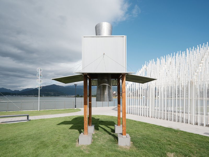 White cubic volume elevated on timber columns with a flat roof beside a vertical rod sculpture