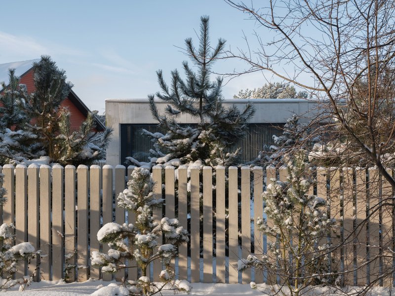 Street view of the concrete volumes behind a vertical timber fence with snow-laden pines