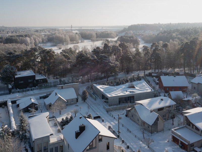 Aerial perspective of the angular roof with parallel skylights surrounded by residential rooftops and a forest in winter