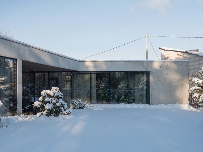 Concrete facade with floor-to-ceiling glazing and timber cladding beside a snow-covered garden under clear winter sky