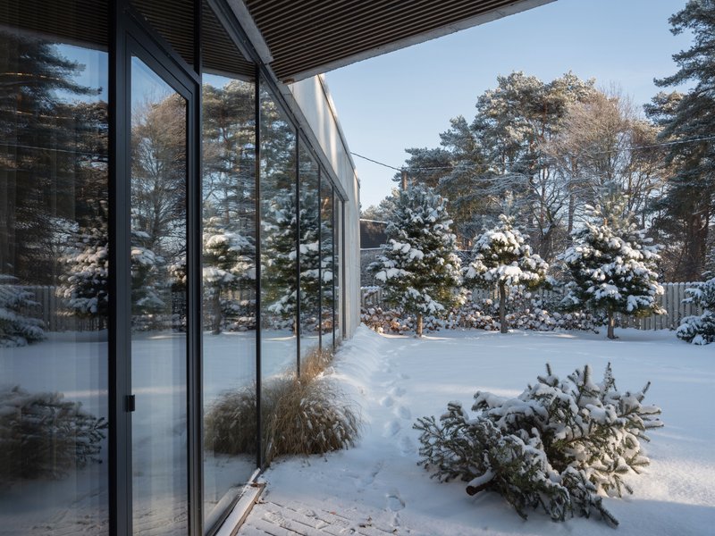 Glass facade with dark frames overlooking a snow-covered garden with planted beds and evergreen shrubs
