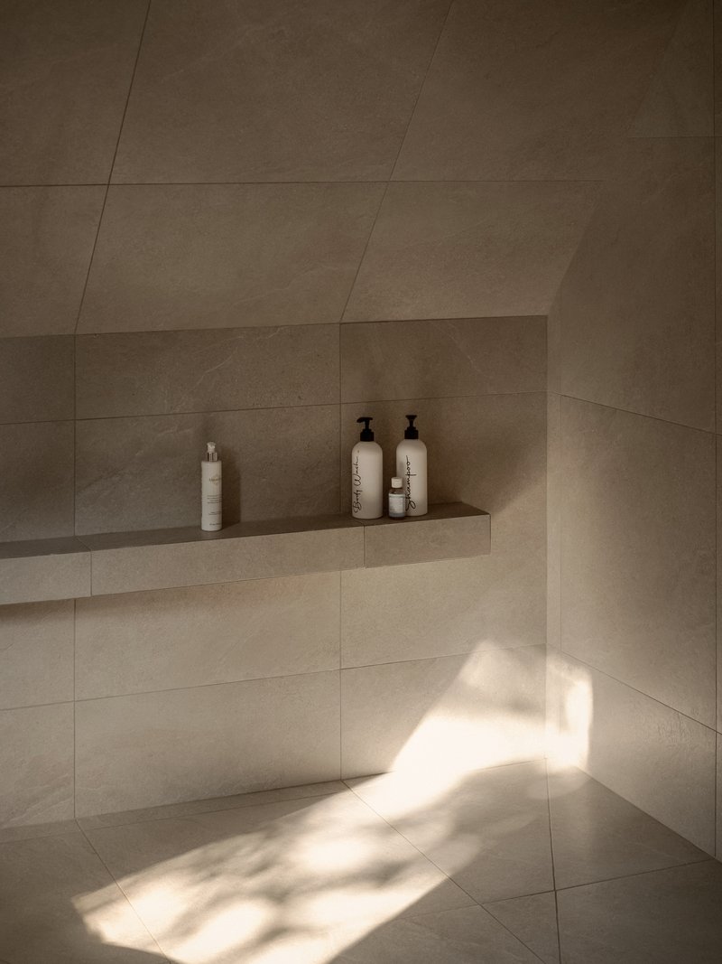Recessed stone tile shower niche with white bottles illuminated by sunlight filtering across the floor