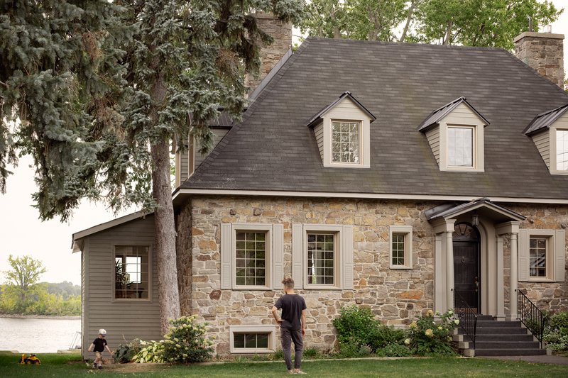 Stone facade with dormer windows and steeply pitched slate roof beside a lawn with figures and waterside trees