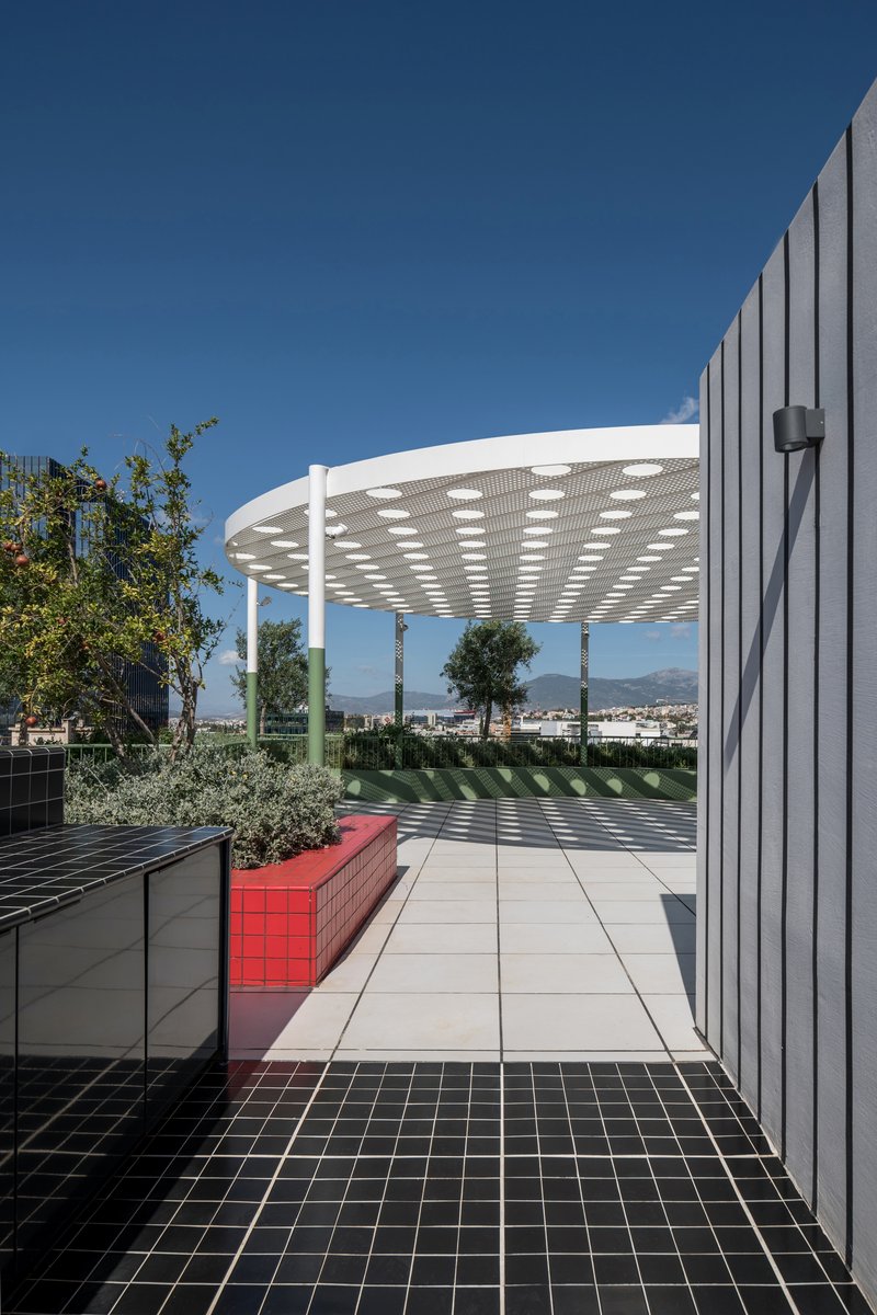 Rooftop terrace with circular perforated canopy, black tile paving, and red bench under clear blue sky