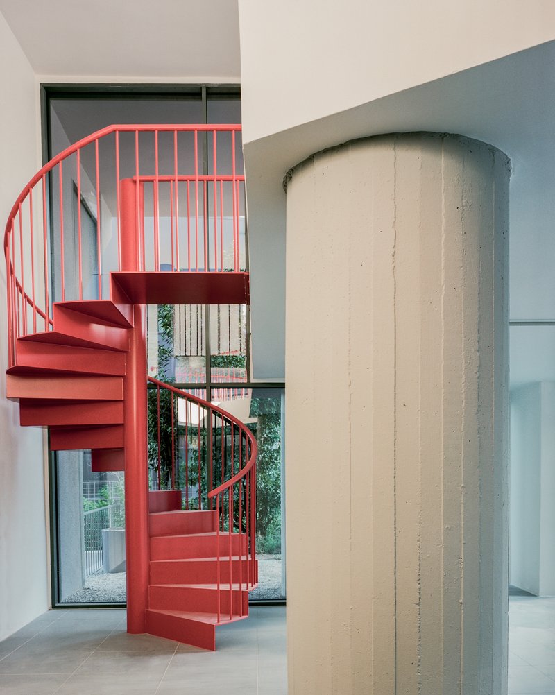 Red spiral staircase beside a cylindrical concrete column with garden views through glazed opening