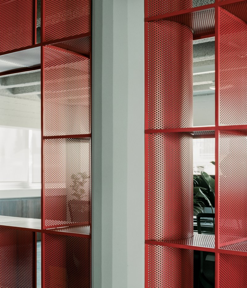 Close-up of red perforated metal shelving units with curved segments reflecting light from workspace beyond
