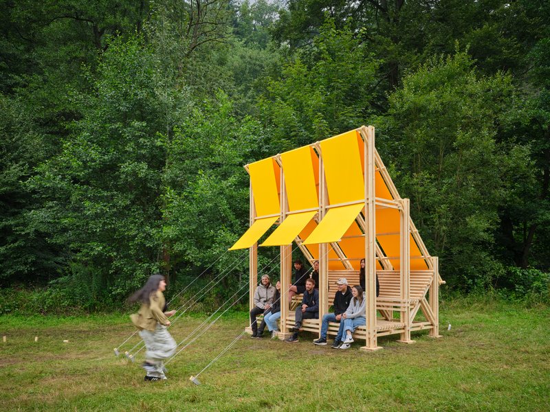 Open timber frame pavilion with yellow fabric canopy sheltering seated visitors in a meadow