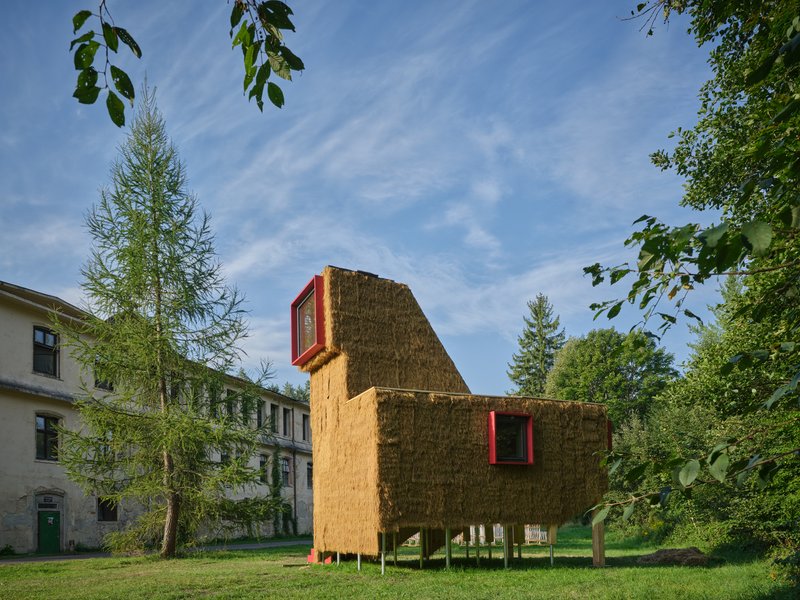 Elevated straw bale pavilion with red window frames on a grassy lawn beside existing institutional buildings