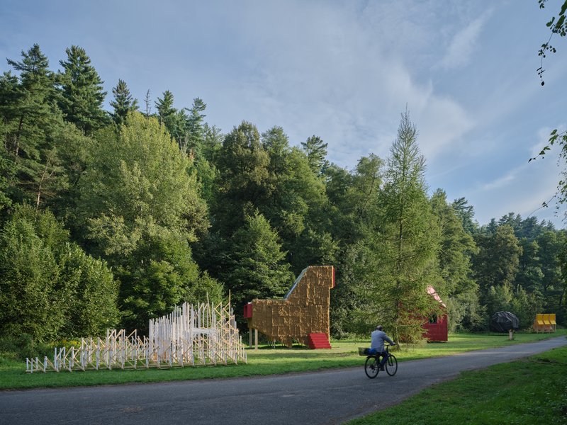 White slatted timber fence enclosure and straw-clad pavilion with a cyclist on the pathway