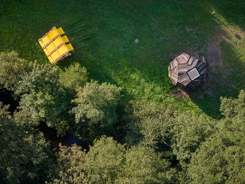 Drone view of yellow pavilion and faceted timber structure on a clearing edged by forest