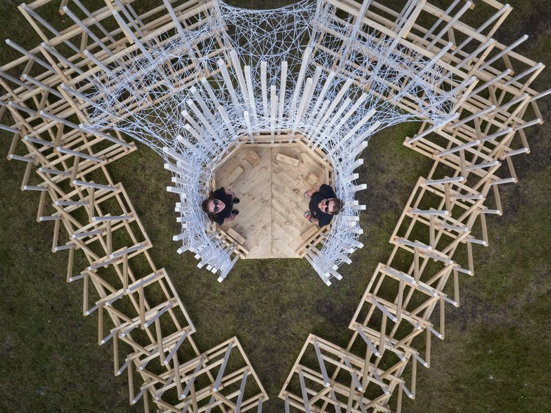 Aerial view of a radial timber pavilion with white lattice canopy and two people standing on the central platform