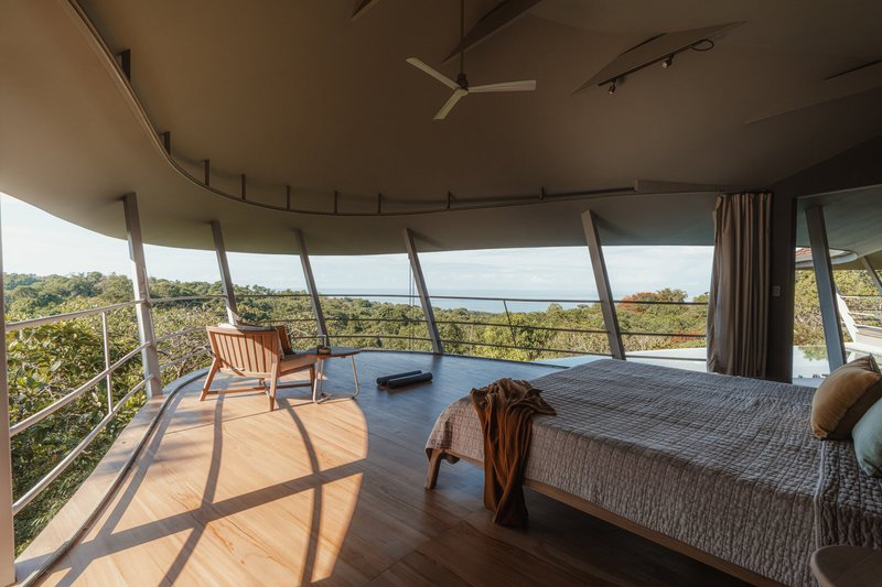 Bedroom interior with the bed positioned toward the curved glass wall and a view of the ocean in the distance