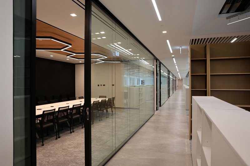 Glass-walled corridor with conference rooms on one side and timber shelving opposite under linear ceiling lights