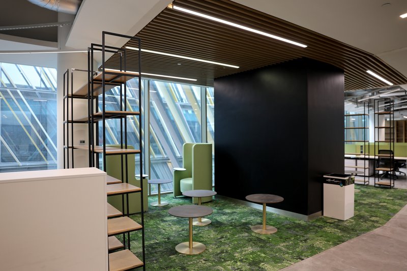 Lounge zone with green upholstered seating pods on grass carpet beside diagonal-braced glazed facade