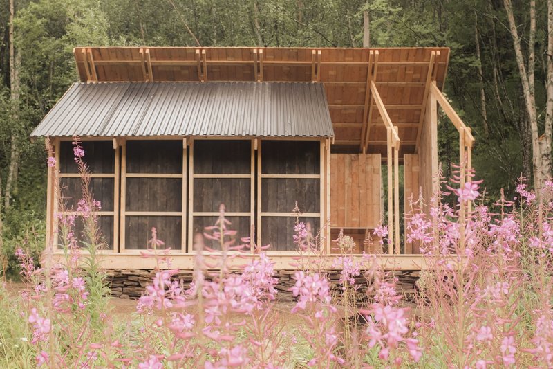 Timber frame structure with corrugated metal roof and screened walls framed by pink wildflowers