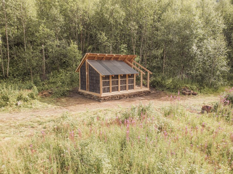 Gabled timber pavilion with stone base and metal roof in a forest clearing