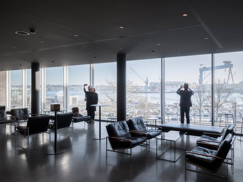 Interior lounge with black leather chairs and floor-to-ceiling windows overlooking industrial waterfront cranes