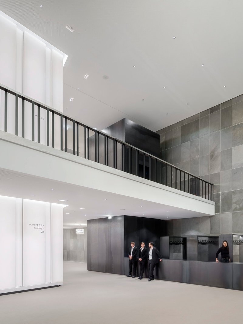 Double-height lobby space with black metal railing balcony and grey tiled wall beside reception desk