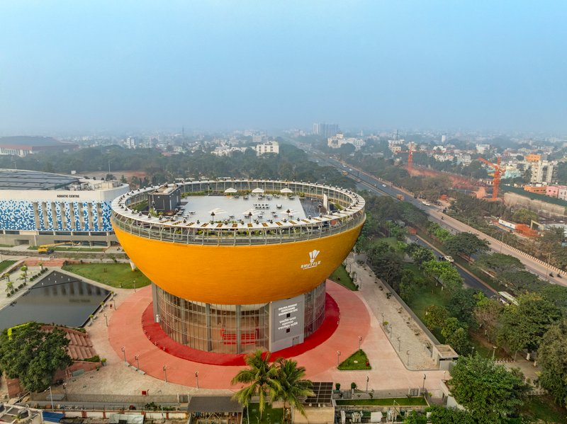 Drone perspective showing the circular rooftop plaza with glazed perimeter colonnade atop the yellow-clad bowl form
