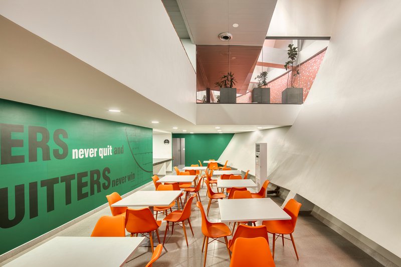 Interior dining area with orange chairs and green wall graphics beneath a skylit mezzanine with planters