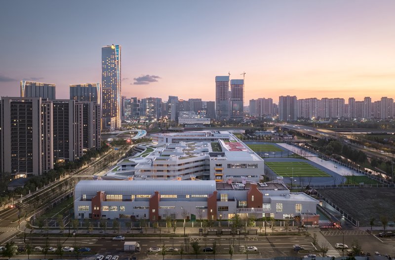 Long view of the low-rise white volume with rooftop sports courts and surrounding residential towers at sunset