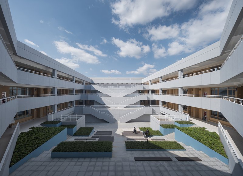 Courtyard framed by stacked white volumes with landscaped beds and a person seated under blue sky