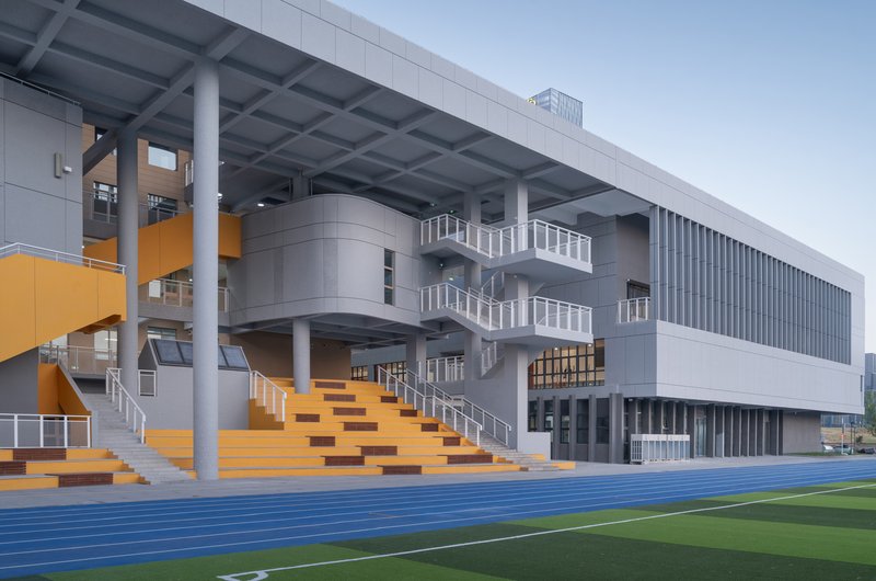 Orange tiered seating stairs beneath deep overhang beside blue athletic track and green sports field