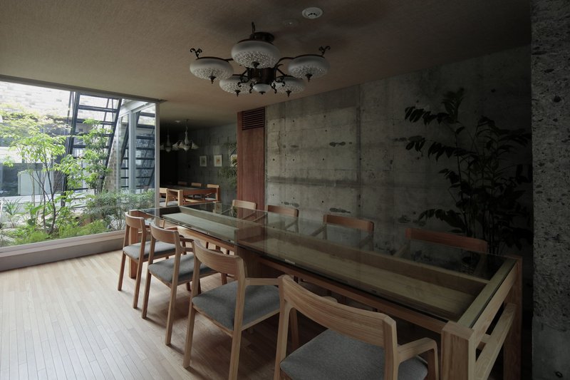Dining room with glass-topped timber table, concrete walls, chandelier, and courtyard garden visible through corner windows
