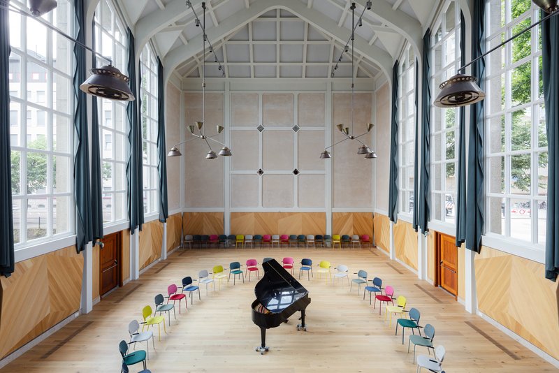 Vaulted performance hall with grand piano surrounded by colorful seating under industrial pendant lights