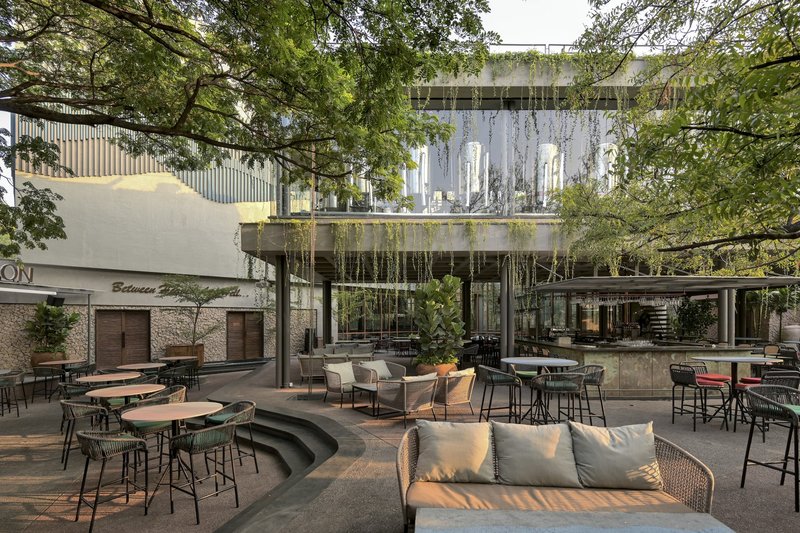 Outdoor courtyard with curved seating platform and cafe tables beneath overhanging planted terraces and surrounding trees