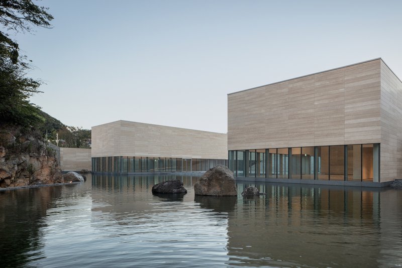 Horizontal stone-clad volumes reflected in a still pond with large boulders at dusk