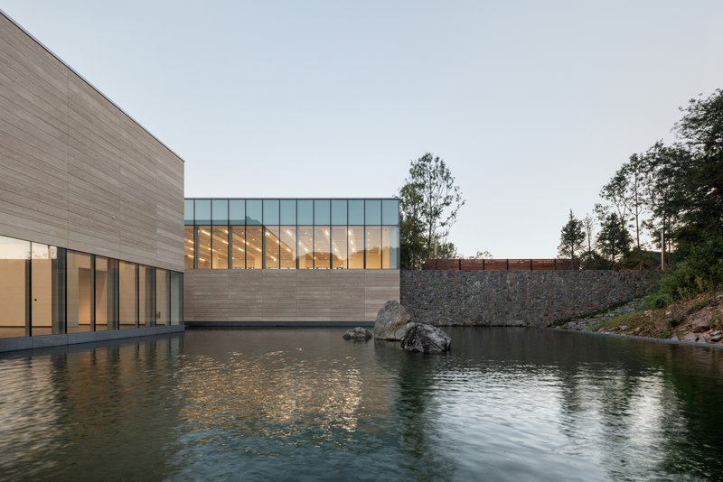 Clerestory glazing above horizontal stone-clad base beside water pool with gabion retaining wall
