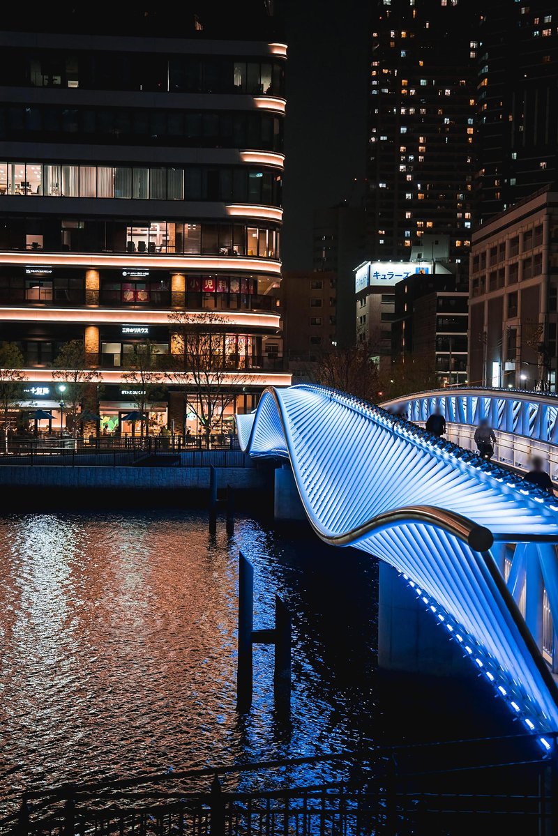 Curved pedestrian bridge with blue illuminated pleated metal canopy reflected in the canal below
