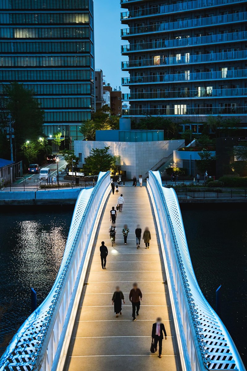 Illuminated pedestrian bridge with perforated steel railings spanning a canal at dusk with people walking