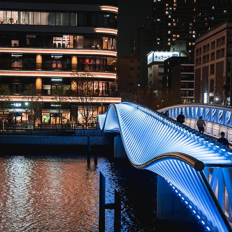 Undulating footbridge with LED-lit ribbed structure crossing dark water beside a commercial building at night