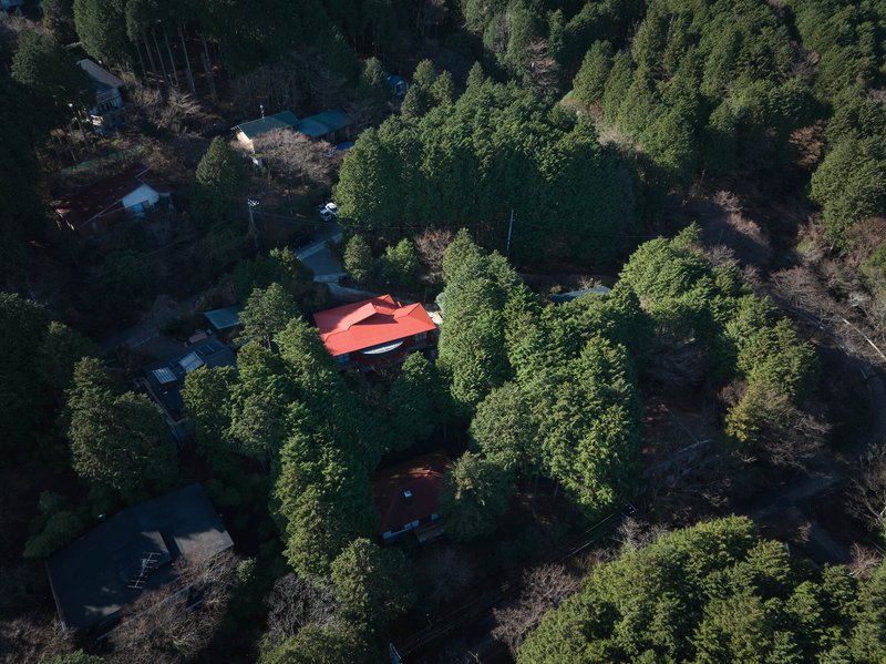 Drone view of the red roof among wooded slopes with scattered neighboring structures visible in the clearing