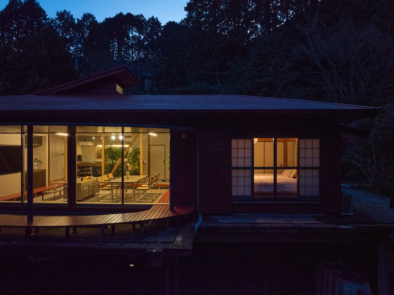 Illuminated living pavilion with glazed walls and timber deck at nightfall beside a dark reflecting pond