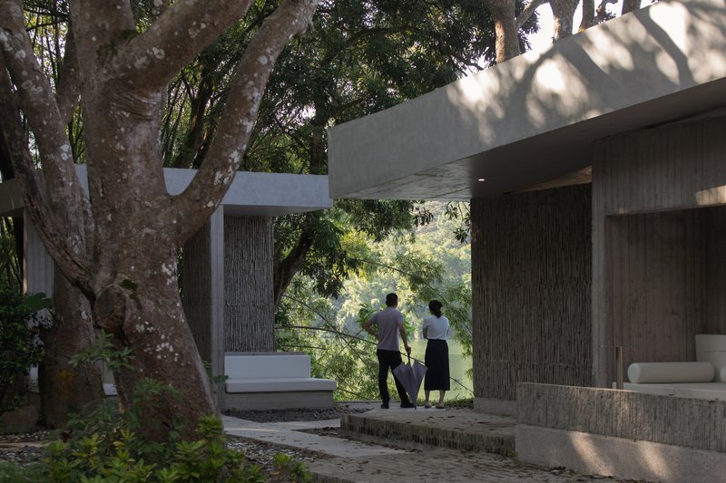 Two figures standing on a covered terrace framed by concrete beams and mature tree canopy