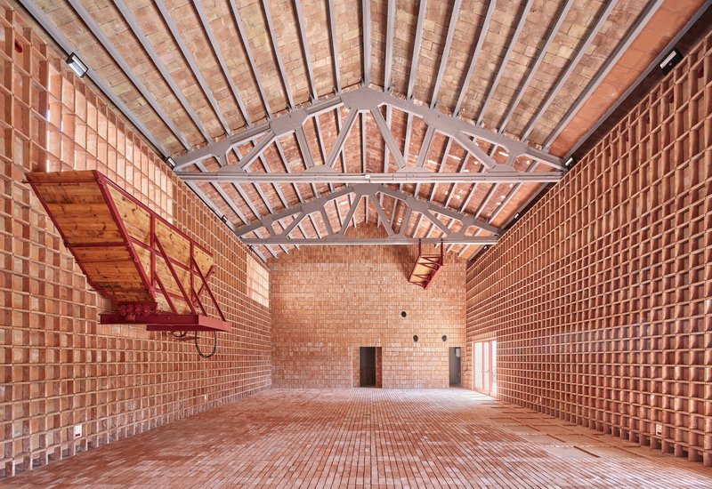 Interior hall with perforated terracotta brick walls, exposed timber trusses and a red steel balcony under diffused daylight