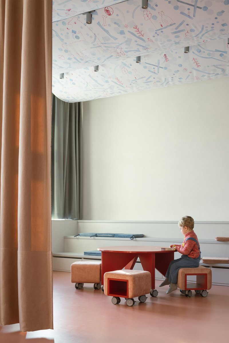 Child seated at red table beneath patterned fabric ceiling with beige and green curtains framing window