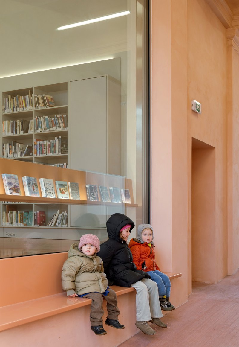Three children in winter jackets seated on pink built-in bench beside glass display case