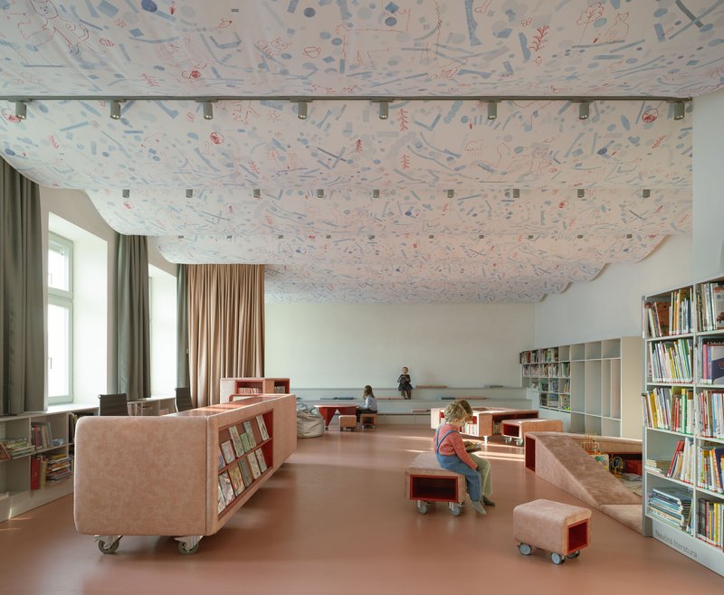 Library interior with patterned fabric ceiling, mobile pink shelving units, and children reading on a terracotta floor