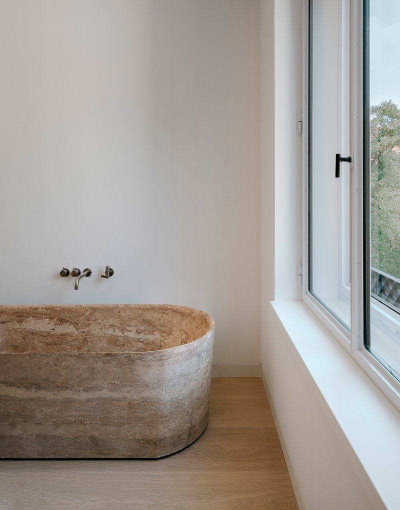 Freestanding concrete bathtub beneath wall-mounted faucets next to a narrow casement window with treetops visible outside